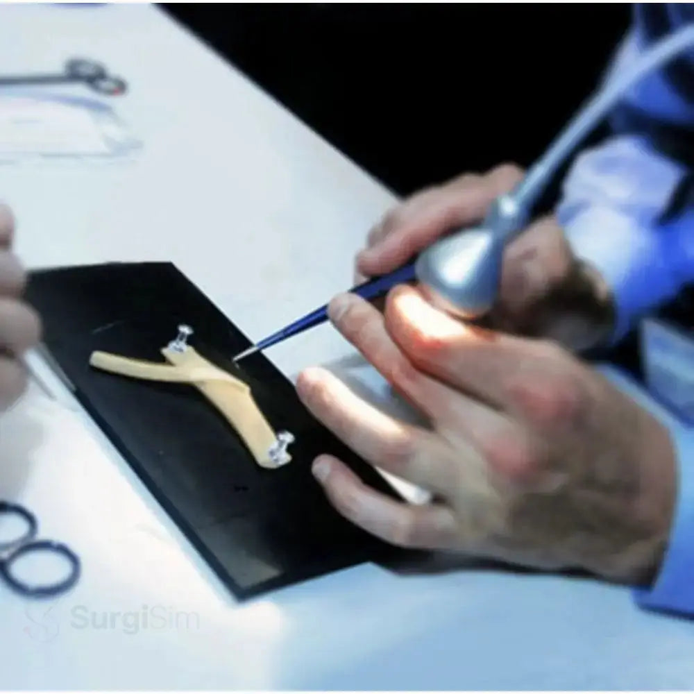 A silver surgical instrument with a blue handle held over a black tissue mounting pad for anastomosis practice